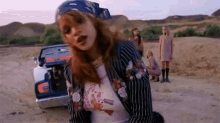 a woman wearing a bandana and a striped jacket is kneeling in the dirt in front of a car .