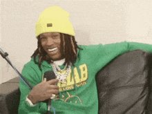 a man wearing a yellow beanie and a green hoodie is sitting on a couch with a microphone .