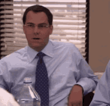 a man in a blue shirt and tie is sitting at a table with a water bottle in front of him .