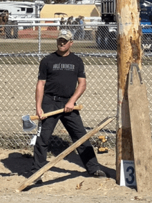 a man wearing a black shirt that says able ebenezer holds a large wooden stick