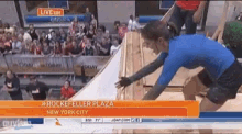 a woman in a blue shirt is jumping over a wall in front of a crowd at rockefeller plaza new york city