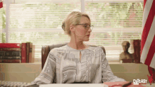 a woman wearing glasses sits at a desk in front of a window with outcast written on the bottom right