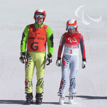 a man and a woman are walking down a snowy slope and the man is wearing a vest that says beijing 2022