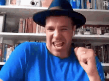 a man wearing a hat and a blue shirt is making a funny face while sitting in front of a bookshelf .