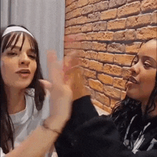 two young women are sitting next to each other in front of a brick wall and talking to each other .