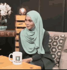 a woman wearing a hijab is sitting on a couch with a cup of coffee on the table .