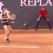 a man in a red shirt is watching a woman play tennis in front of a sign that says replay