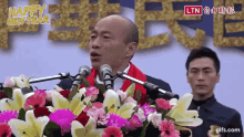 a man is giving a speech at a podium with flowers in front of him and the words happy new year written above him