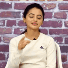 a woman in a white adidas shirt is sitting in front of a brick wall and making a funny face .