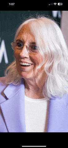 a woman wearing glasses and a purple jacket is smiling at the camera with the time of 1:07