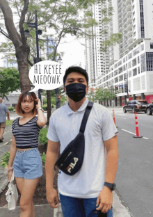 a man wearing a mask stands next to a woman wearing shorts and a fila waist bag