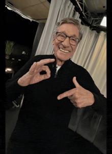 an older man wearing glasses and a black sweater is making a peace sign