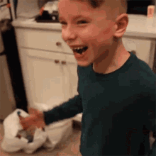 a young boy in a green shirt is laughing in a kitchen while standing next to a trash can .