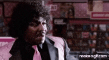 a man with curly hair and a pink tie is sitting in front of a shelf .