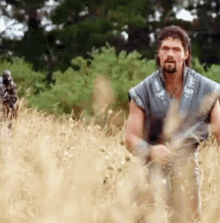 a man with a beard is walking through a field of tall grass with a horse in the background .