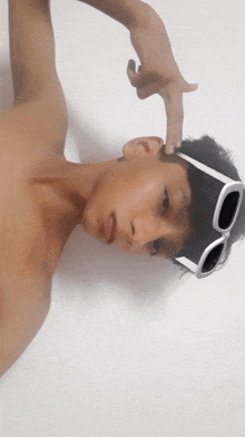 a shirtless boy wearing sunglasses is pointing his finger at his head