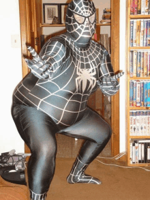 a man in a spider man costume is squatting in front of a bookshelf filled with dvds