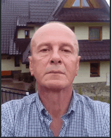 a bald man in a plaid shirt takes a selfie in front of a house