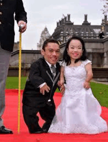 a man and a woman are standing next to each other on a red carpet . the woman is wearing a wedding dress .