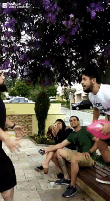 a group of people sitting on a bench under a tree with purple flowers and a facebook video coming up