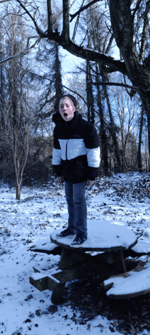 a boy in a black and white jacket stands on a snowy picnic table