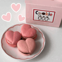 three heart shaped cookies are on a plate next to a pink box that says cookie