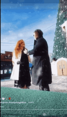 a man in a black coat is standing next to a woman with red hair in front of a christmas tree