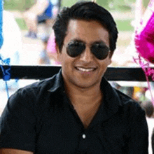 a man wearing sunglasses and a black shirt is smiling and sitting in front of balloons .