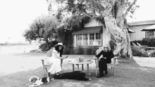 a black and white photo of two women sitting in chairs with their dogs in front of a house .