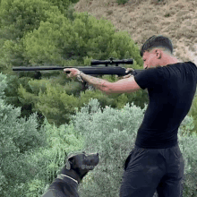 a man in a black shirt is holding a rifle while a dog looks on