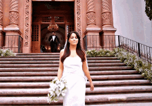 a woman in a white dress stands on a set of stairs with a man in a tuxedo behind her