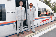 two men in suits are standing in front of a train that says catch on it