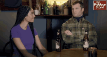 a man and a woman are sitting at a table with bottles of beer and a sign that says produce stand