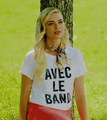 a woman is sitting under a tree wearing a white t-shirt with the words `` avec le banauc '' on it .