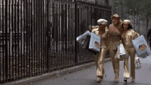 three women in gold jumpsuits are walking down a sidewalk carrying shopping bags including one that has a picture of a woman on it