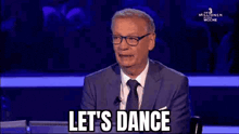 a man in a suit and tie is saying `` let 's dance '' while sitting at a desk .