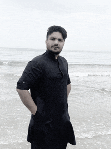 a man in a black shirt is standing on a beach with his hands in his pockets