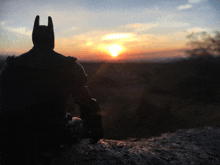 a silhouette of batman sitting on a rock at sunset