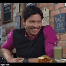 a man in a pink shirt is sitting at a table with a plate of food and a drink .