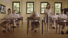 a group of students are sitting at desks in a classroom while a teacher stands in front of them .