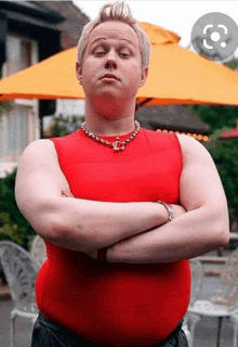 a man wearing a red tank top and a necklace is standing with his arms crossed