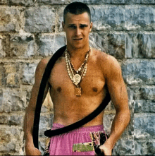 a shirtless man wearing a gold necklace and pink shorts is standing in front of a brick wall