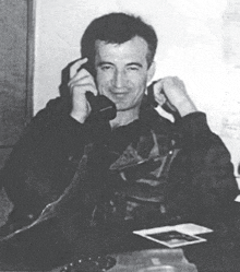 a man in a leather jacket is sitting at a desk talking on a phone .