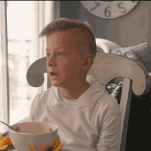 a young boy sits in a chair with a bowl of cereal in front of a clock that says 265