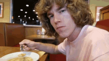 a young man with curly hair is sitting at a table with a plate of food and a fork in his hand .