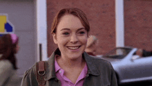 a young woman wearing a pink shirt and a green jacket is smiling in front of a brick building .