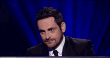 a man in a suit and tie is sitting in front of a blue curtain