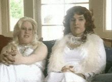 two women are sitting on a couch and one is wearing a white dress