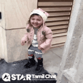 a little girl wearing a pink jacket and a hat is smiling in front of a sign that says tamil chat