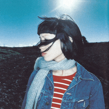 a woman wearing a scarf and a denim jacket is standing in a field